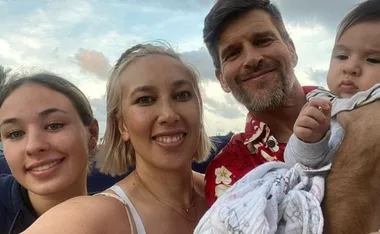 Family selfie with three adults and a baby smiling outdoors, under a partly cloudy sky.