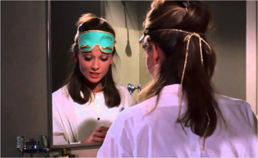 Woman in white robe and teal sleep mask looks into a mirror, reflecting her thoughtful expression.