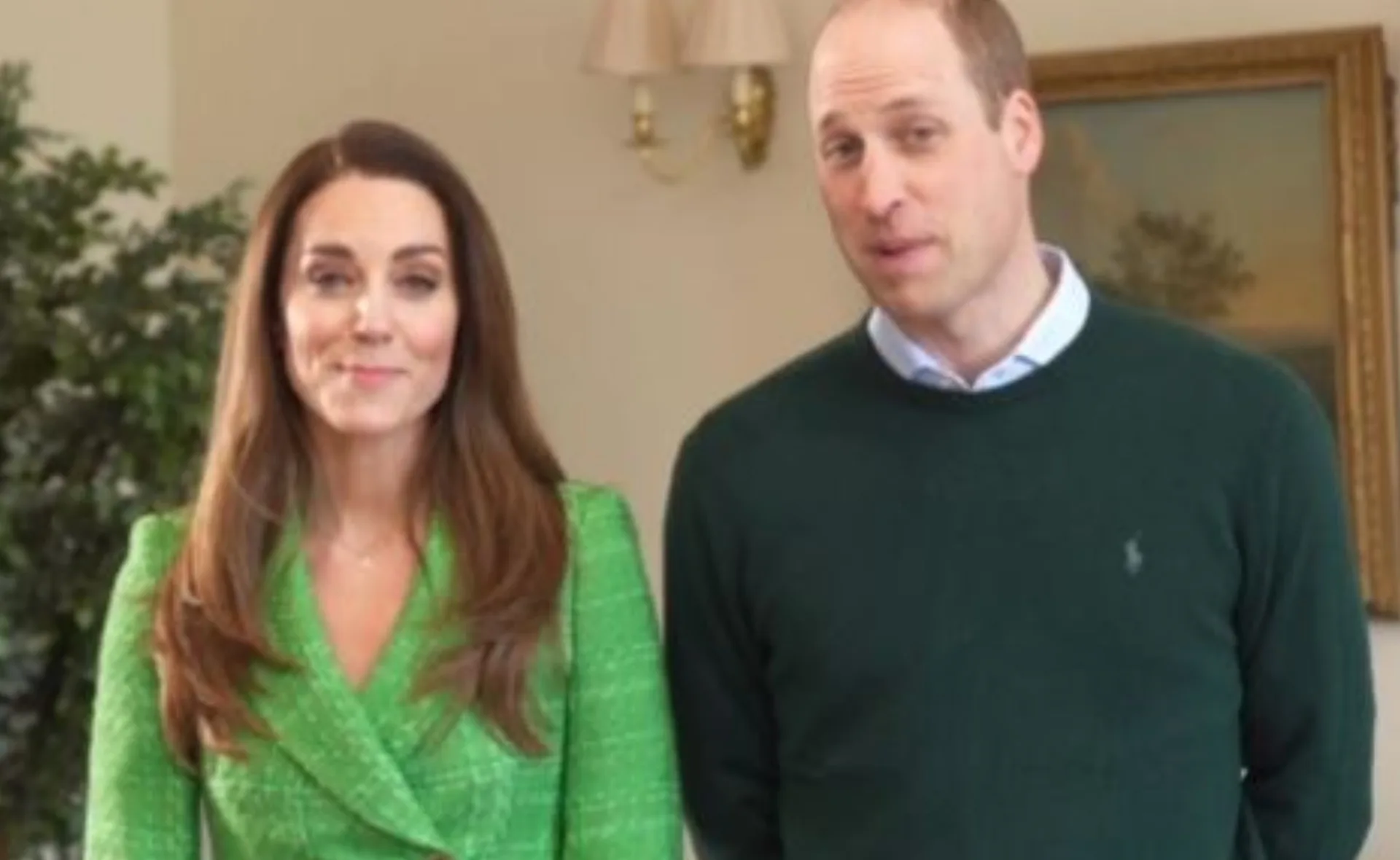 A woman in a green outfit and a man in a green sweater stand together indoors, smiling at the camera.