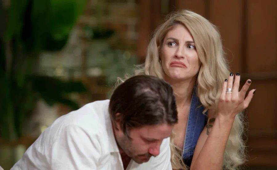A woman with blonde hair looks displeased, while a man with dark hair leans forward. They appear to be indoors.