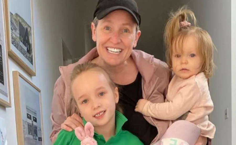 A person smiling with two young children, one wearing green holding a toy, the other in pink being held.