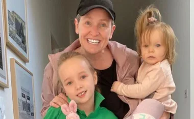 A person smiling with two young children, one wearing green holding a toy, the other in pink being held.