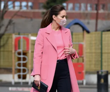 A woman in a pink coat and mask walks outdoors, holding a black clutch with a playground in the background.