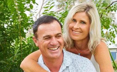 A couple smiling outdoors, surrounded by green foliage.