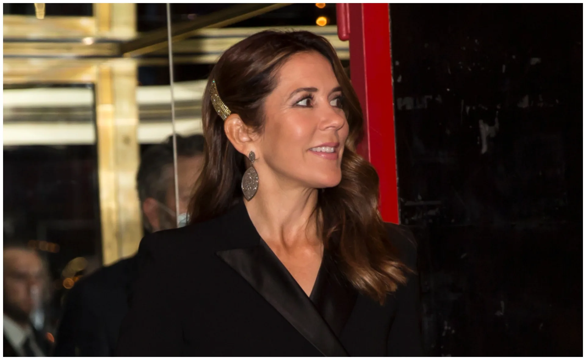 A woman with wavy brown hair smiles, wearing large oval earrings, a black suit, and a gold hair accessory.
