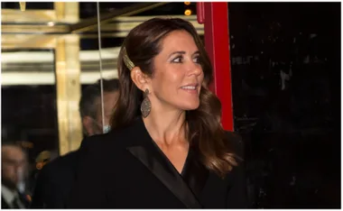 A woman with wavy brown hair smiles, wearing large oval earrings, a black suit, and a gold hair accessory.