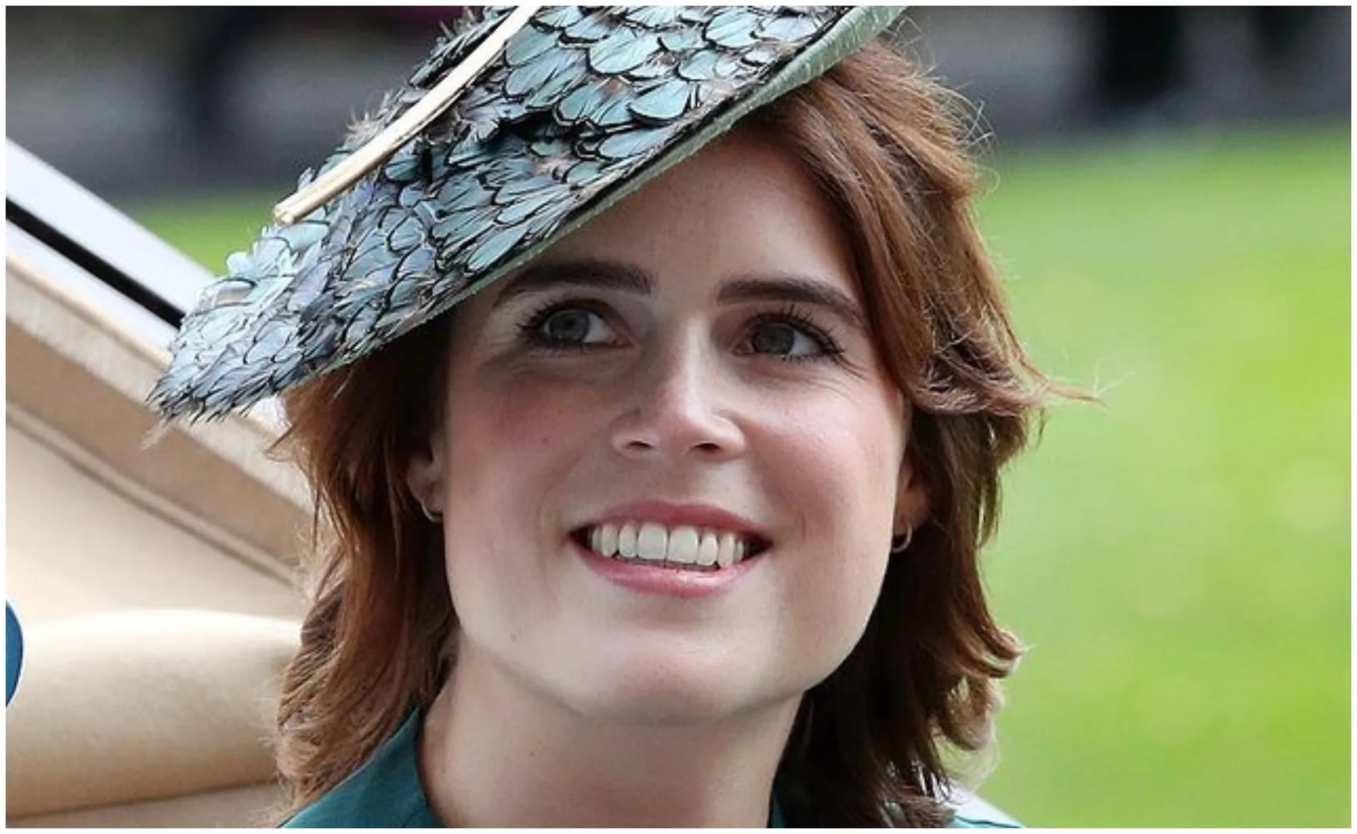 A woman smiling, wearing a feathered hat, outdoors with a blurred green background.