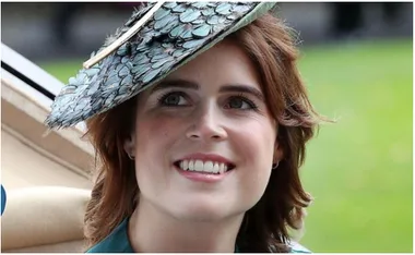 A woman smiling, wearing a feathered hat, outdoors with a blurred green background.
