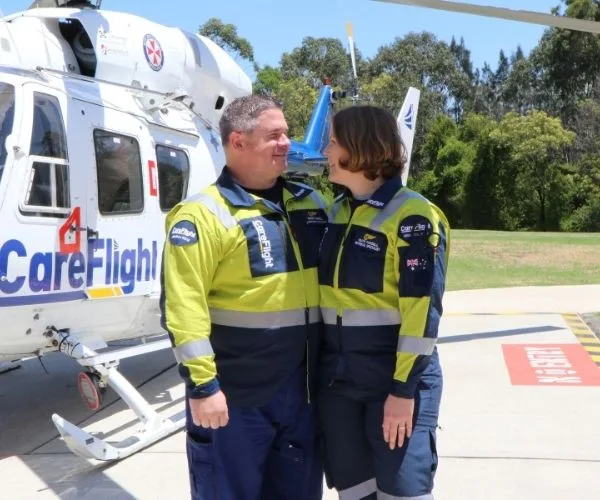 A smiling couple in care flight uniforms stands next to a rescue helicopter on a sunny day.