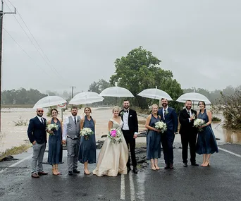 Bride, groom, and wedding party pose with umbrellas on a flood-damaged road.
