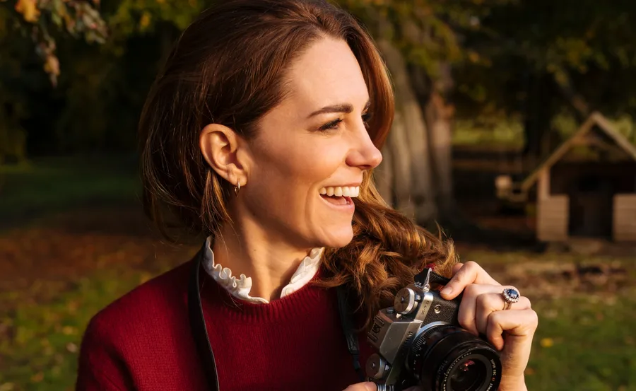 A woman smiling while holding a camera, standing outdoors with trees in the background.