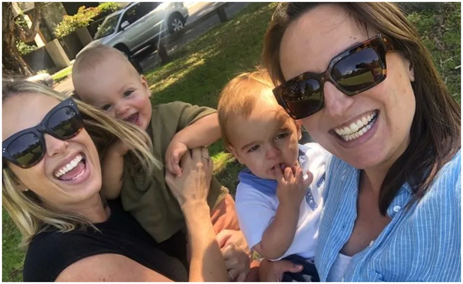 Two women smiling with two toddlers, one woman holding a child in a park setting on a sunny day.