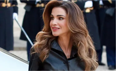 A person with long hair wearing a black leather outfit, smiling with a blurred background of people in uniforms.
