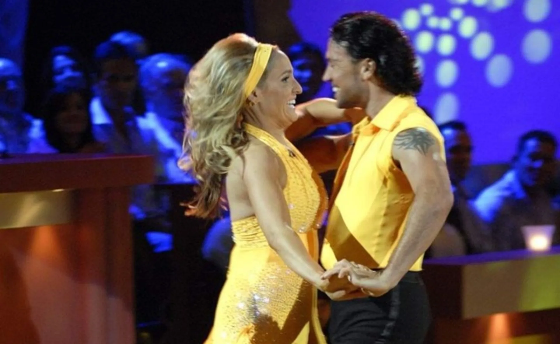 A couple in yellow outfits dancing energetically on a stage with an audience in the background.