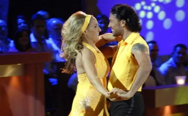 A couple in yellow outfits dancing energetically on a stage with an audience in the background.