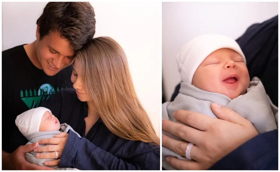A couple lovingly holding their newborn wrapped in a blanket; baby smiling with eyes closed, wearing a white hat.