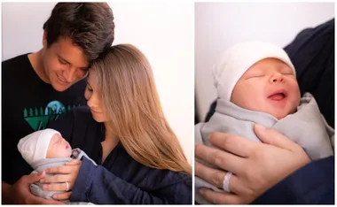 A couple lovingly holding their newborn wrapped in a blanket; baby smiling with eyes closed, wearing a white hat.