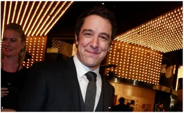 A man in a suit smiles under bright marquee lights at a formal event, with a woman in the background.
