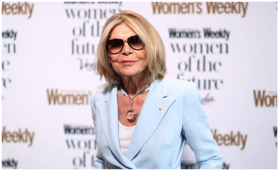 Blonde woman in sunglasses and blue blazer at an event, posing confidently in front of branded backdrop.