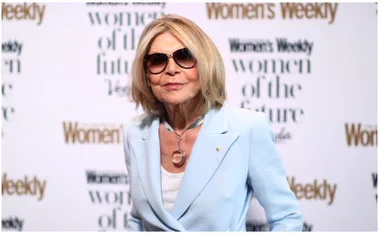 Blonde woman in sunglasses and blue blazer at an event, posing confidently in front of branded backdrop.