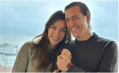 A smiling couple with one displaying a ring, posing in front of a cityscape with a cloudy sky.