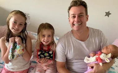 A smiling man with three children; two girls holding toys, and a baby asleep in his arms with a stuffed bunny.