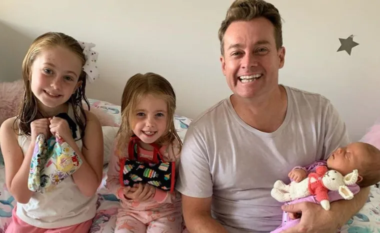A smiling man with three children; two girls holding toys, and a baby asleep in his arms with a stuffed bunny.