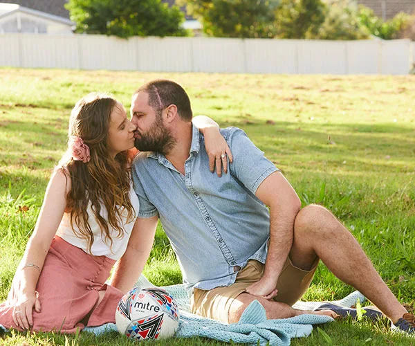 Couple sitting on grass kissing, with a soccer ball and blanket nearby in a sunny park setting.