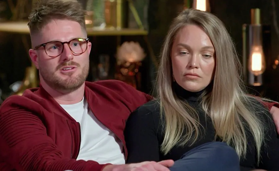 Couple sitting together on a couch, man in glasses and red jacket, woman with long blonde hair, neutral expressions.