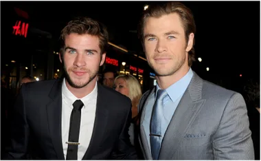 Two men in suits smiling at an event; night-time setting with stores in the background.