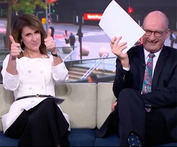 Two people sitting on a couch, one giving thumbs up and the other holding papers, both are smiling.