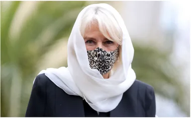 Woman in a white headscarf and leopard-print mask, wearing a dark coat, with a blurred outdoor background.