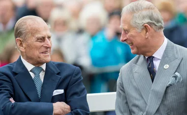 Two men in suits share a laugh outdoors, surrounded by a blurred crowd.