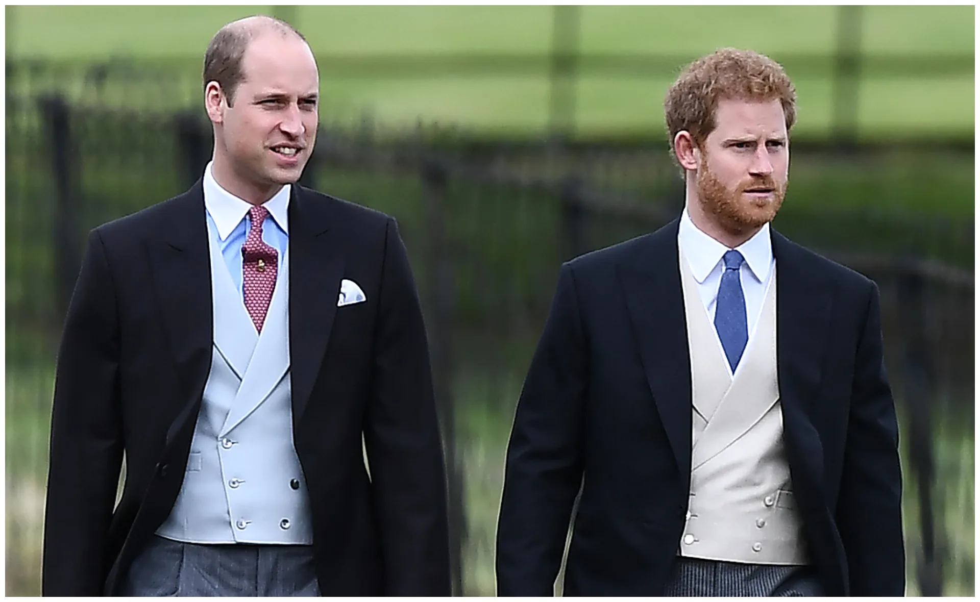 Two men in formal suits walking outside with a blurred greenery background.