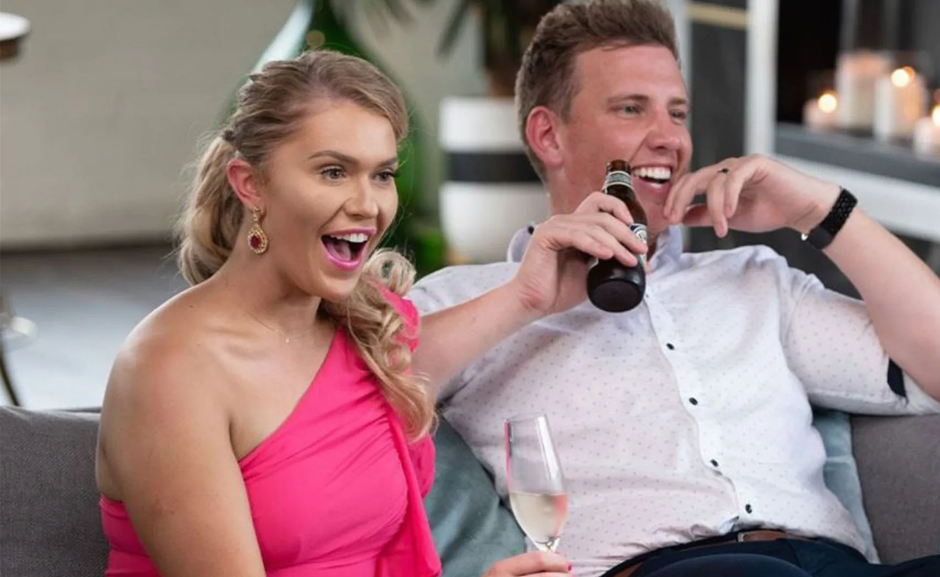 A couple sitting on a couch, smiling and enjoying drinks; one holding a beer, the other a glass of wine.