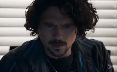 A man with curly hair and facial hair looks down, focused, in a dimly lit room with blinds in the background.