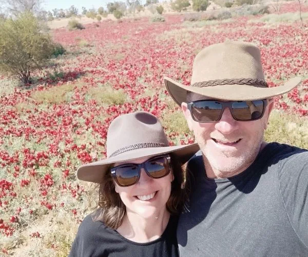 Couple in hats and sunglasses smiling in a field of red wildflowers under a clear blue sky.