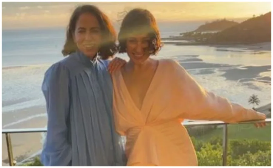 Two people in pastel outfits pose on a balcony overlooking a coastal landscape at sunset.