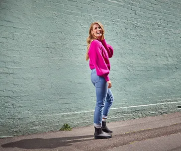 Person in a pink sweater, jeans, and black ankle boots standing against a light green wall, looking back and smiling.
