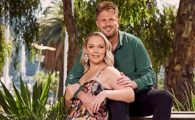 Couple sitting outdoors, smiling and embracing, with greenery in the background.