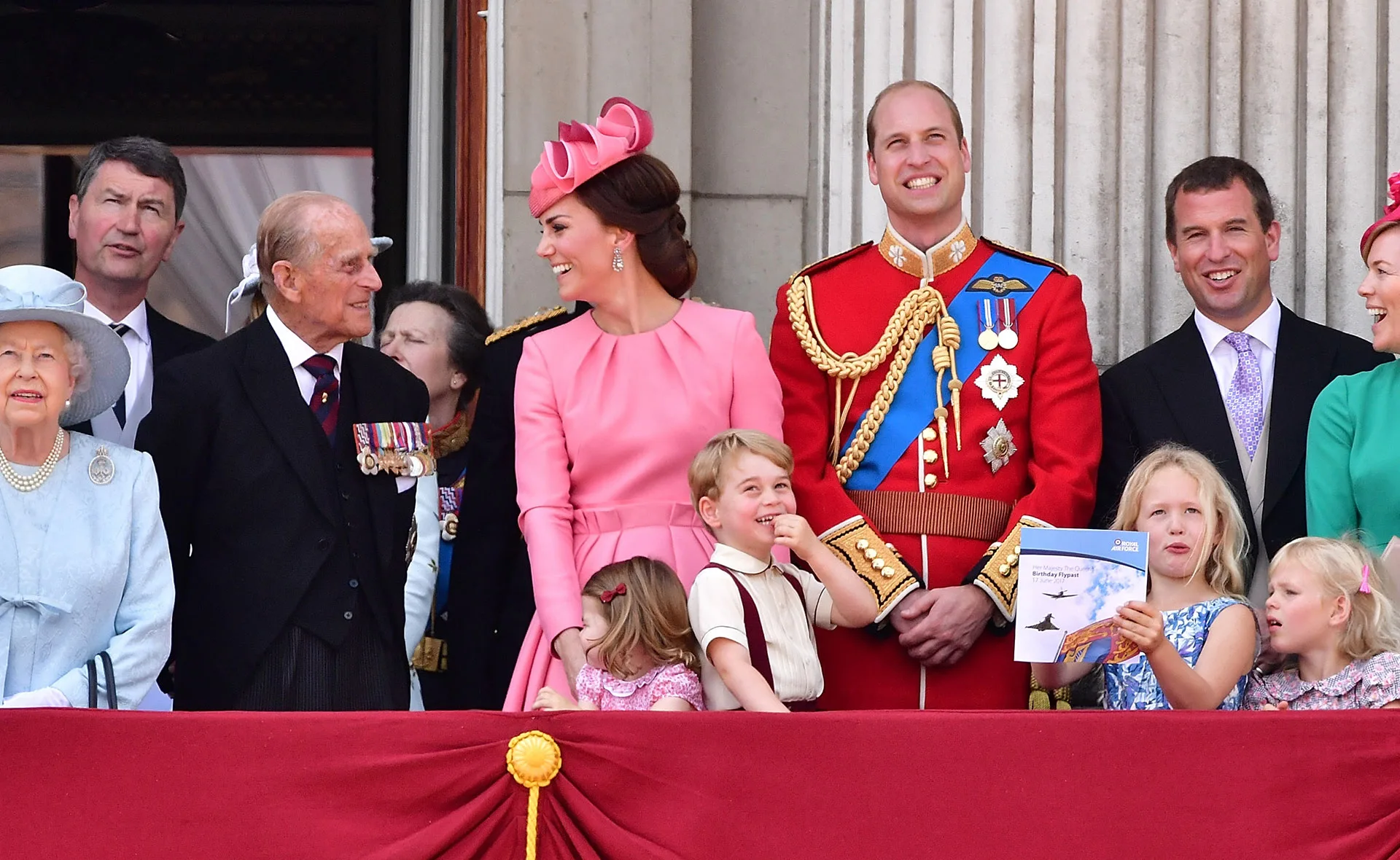 A family gathering on a balcony, adults smiling, children holding a pamphlet, dressed in formal attire with medals displayed.