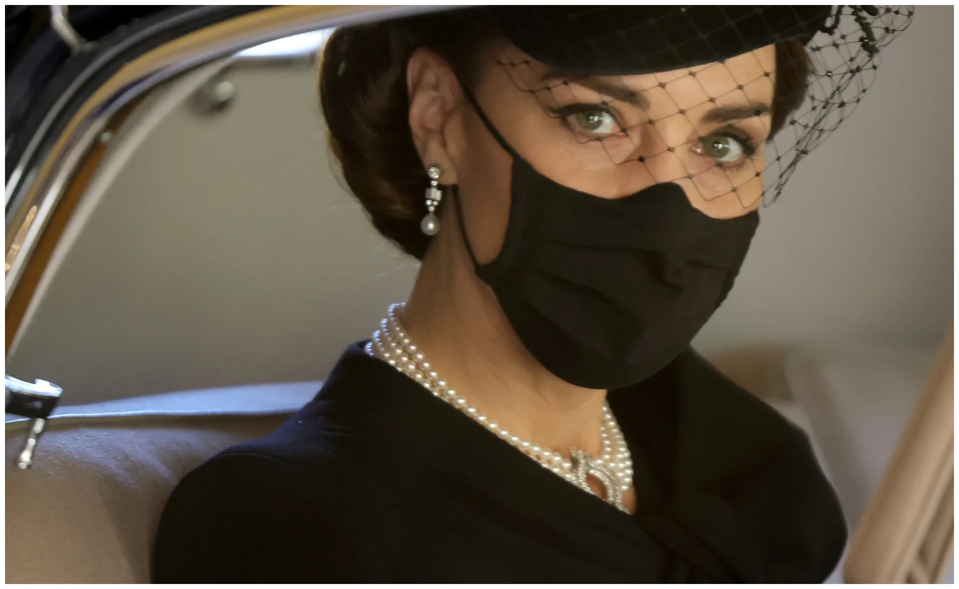 Woman in black attire with pearls and a black hat, wearing a face mask, sitting in a car.