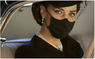 Woman in black attire with pearls and a black hat, wearing a face mask, sitting in a car.