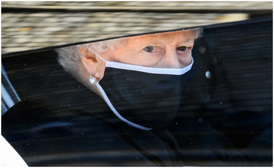 A person wearing a black face mask and pearl earrings, viewed through a car window.