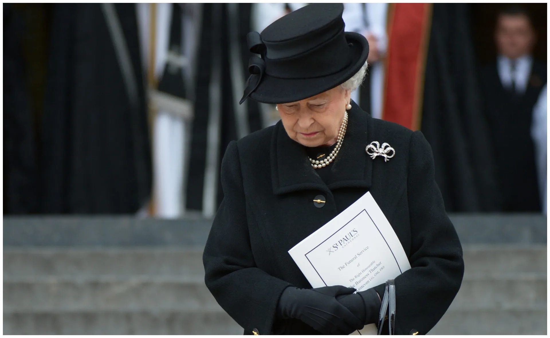 Elderly woman in black attire, holding a funeral program, head bowed in solemn reflection.