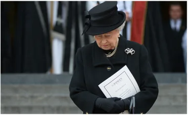 Elderly woman in black attire, holding a funeral program, head bowed in solemn reflection.