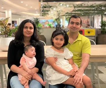 A family of four, parents and two young children, sitting at a table in a bright indoor setting.