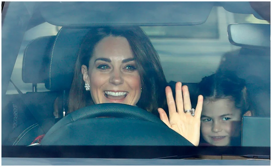 Woman smiling and waving from car, child seated next to her, seen through the windshield.