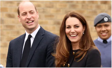 Prince William and Kate Middleton smiling outdoors, with an air cadet in the background.