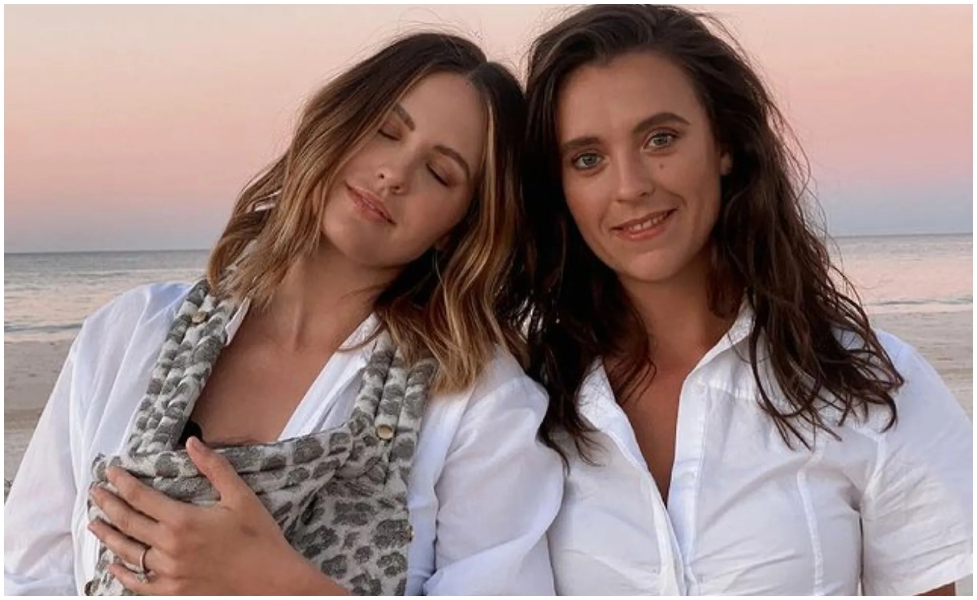 Two women in white shirts stand on a beach at sunset, one resting her head on the other's shoulder.